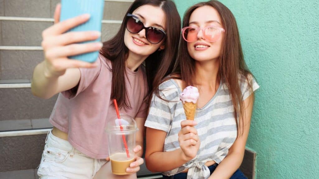 Two girls with an ice cream and an iced coffee take a snapshot on their cellphone.